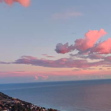Eine friedliche Küstenlandschaft bei Sonnenuntergang, mit zartrosa Wolken, die über den Himmel verstreut sind, ruhigem, blauem Meerwasser und einem kleinen Küstenort, der sich an die Küste schmiegt.
