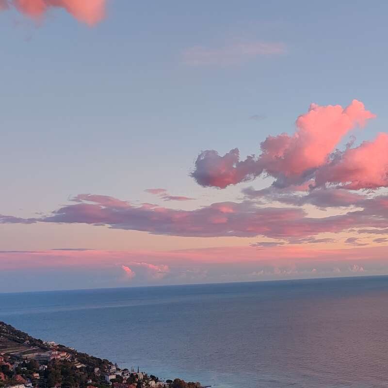 Eine friedliche Küstenlandschaft bei Sonnenuntergang, mit zartrosa Wolken, die über den Himmel verstreut sind, ruhigem, blauem Meerwasser und einem kleinen Küstenort, der sich an die Küste schmiegt.