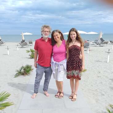 A imagem mostra uma família de três pessoas em pé em uma praia, com o homem vestindo uma camisa vermelha e calça cinza, a mulher com uma blusa rosa e shorts brancos e a menina com um vestido marrom.