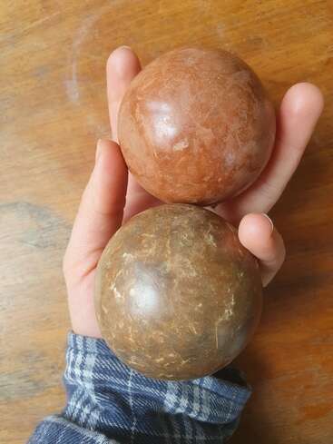 Uma mão de pele clara segura duas bolas marrons, uma acima da outra, com um manguito xadrez azul e branco visível no pulso, contra uma superfície de madeira.