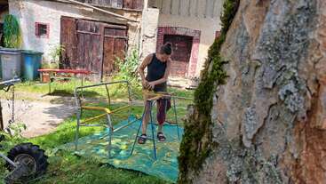 Uma pessoa está em uma lona verde ao ar livre, trabalhando em madeira com ferramentas. Prédios antigos, uma árvore e vários itens de jardim cercam a área sombreada e rústica.
