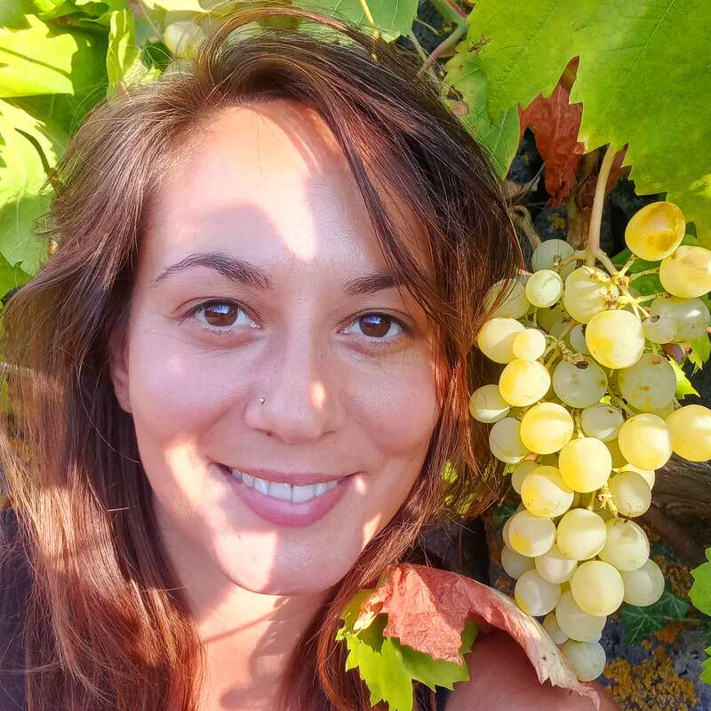 The image depicts a woman with long brown hair, smiling at the camera, surrounded by green leaves and white grapes on a vine.