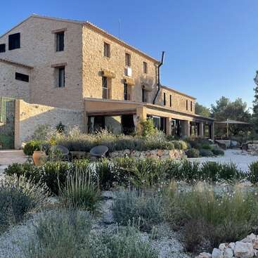 A rustic stone house with a tiled roof sits amid a Mediterranean garden. Outdoor seating, lush greenery, and clear blue skies create a peaceful atmosphere.