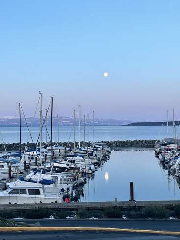 Ein ruhiger Yachthafen in der Abenddämmerung, die Boote liegen ordentlich am Kai. Der Vollmond geht über den fernen Bergen auf, sein Spiegelbild glitzert auf dem stillen, spiegelglatten Wasser. Eine friedliche Szene.