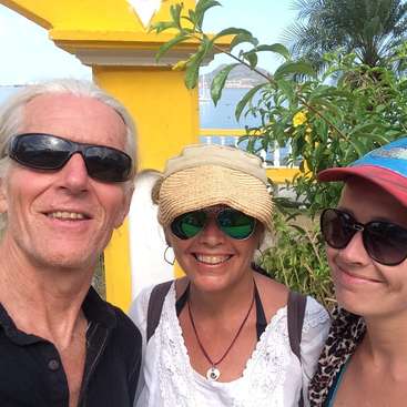 The image shows three people posing for a selfie in front of a yellow building with a body of water and a mountain in the background.