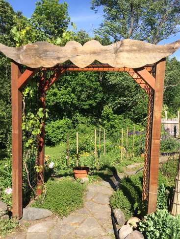 The image depicts a serene garden scene, featuring a wooden arbor with a scalloped top, a stone pathway, and lush greenery, set against a backdrop of trees and a clear blue sky.