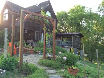 The image depicts a house with a garden and a wooden arbor, featuring a stone pathway, potted plants, and a porch with a screened-in section.