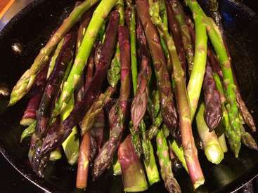 The image depicts a cast iron skillet filled with asparagus spears, showcasing a vibrant display of green and purple hues.