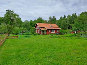 L'image représente une charmante maison rouge avec une pelouse verdoyante, entourée d'arbres, avec un but de football et un trampoline au premier plan.