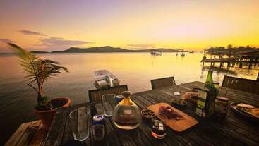 Un coucher de soleil serein sur un lac calme, avec une table en bois pour les boissons et les snacks, des bateaux sur l'eau et des montagnes lointaines créant une ambiance paisible.