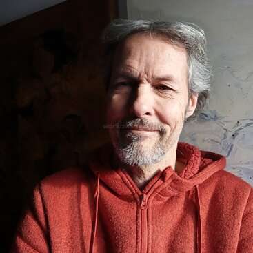 A middle-aged man with gray hair and a short beard is smiling gently, wearing a red hoodie. The background is dark and partially illuminated.