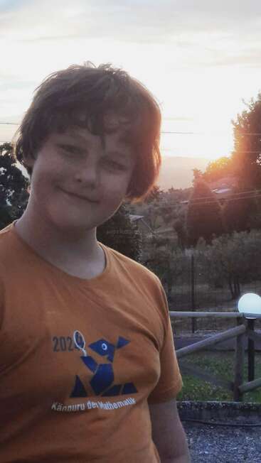 Un garçon souriant, aux cheveux ébouriffés, se tient dehors au coucher du soleil, vêtu d'un t-shirt orange portant l'inscription « Känguru der Mathematik 2021 ». On aperçoit des arbres et une clôture derrière lui.