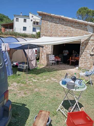 Un campement rustique en plein air, composé d'une tente, d'un auvent, de provisions et d'ustensiles, est installé devant un bâtiment en pierre. Une personne se tient debout à l'intérieur du garage, dont la porte est ouverte.