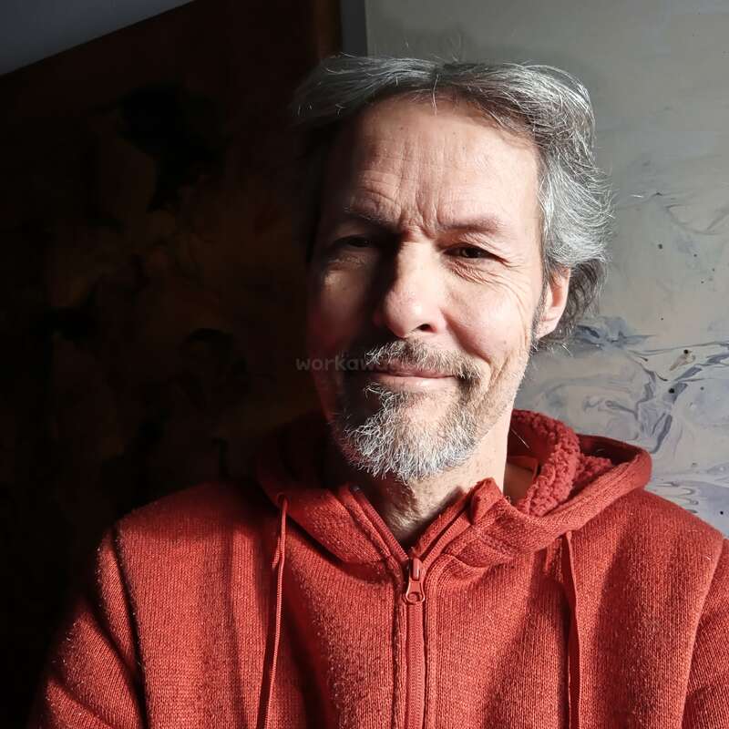 A middle-aged man with gray hair and a short beard is smiling gently, wearing a red hoodie. The background is dark and partially illuminated.