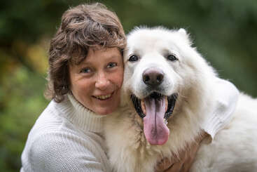 The image depicts a woman with short brown hair, wearing a white sweater, embracing a large white dog with its tongue out, set against a blurred outdoor background.