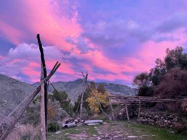 Postes de madera y construcciones rústicas se alzan sobre un terreno rocoso, rodeadas de matorrales y colinas. El cielo resplandece con espectaculares nubes rosadas y moradas al atardecer, creando una atmósfera tranquila.