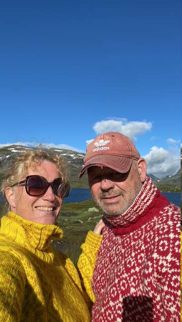Ein lächelndes Paar in bunten Strickpullovern genießt einen sonnigen Tag im Freien; Berge, ein See und blauer Himmel bilden den schönen landschaftlichen Hintergrund hinter ihnen.