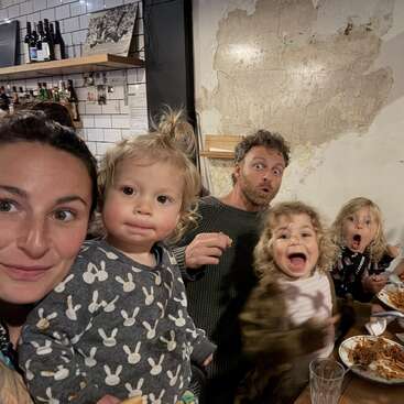 A family of five, including young children, poses for a lively, candid photo at a restaurant table, everyone making silly faces and enjoying their meal together.