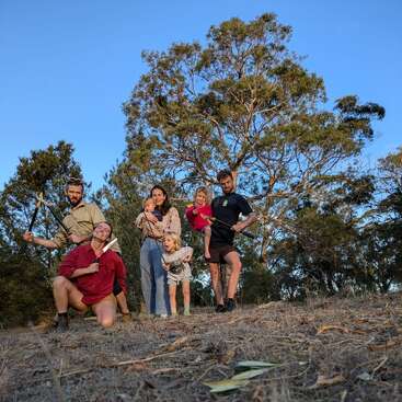 Six people, including children, pose outdoors in a grassy area with trees behind them. Some adults hold sticks or tools, and everyone looks happy and playful.