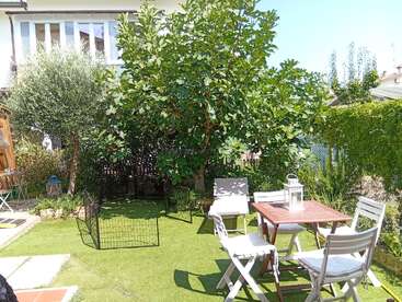 Um jardim ensolarado com árvores verdes exuberantes, uma mesa de madeira e quatro cadeiras brancas, decoração com lanternas externas e uma pequena área cercada com grama artificial.