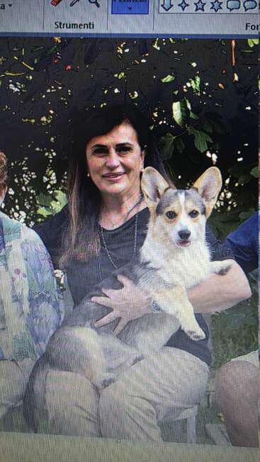 Une femme est assise à l'extérieur, souriante, et tient un adorable chien corgi sur ses genoux. Il y a des arbres en arrière-plan et d'autres personnes à proximité.