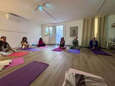 Six femmes sont assises sur des tapis de yoga, les jambes croisées, dans une pièce spacieuse et bien éclairée. L'atmosphère est calme et détendue, suggérant une séance de méditation ou de yoga en groupe.