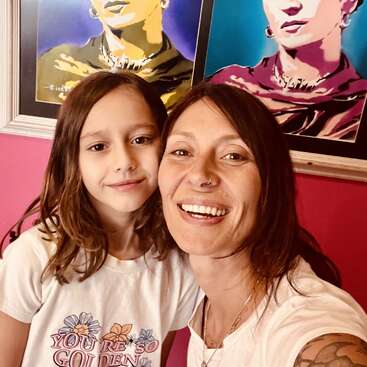 Une femme et un enfant sourient ensemble pour un selfie devant des portraits pop-art colorés sur un mur rose. La chemise de l'enfant porte l'inscription "You're So Golden".