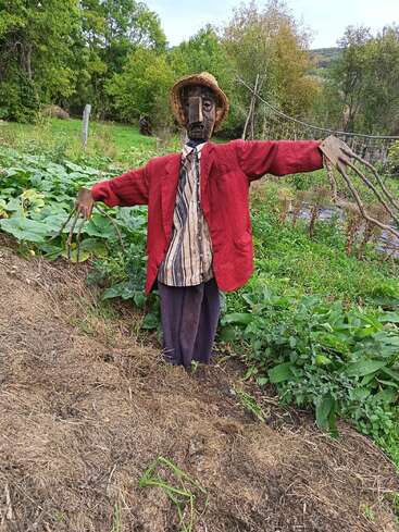 A rustic scarecrow stands in a lush garden, dressed in a red jacket, striped shirt, tie, hat, and wooden hands, surrounded by greenery and trees.