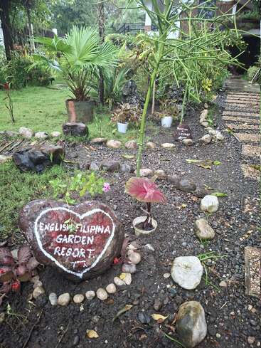 L'entrée d'un centre de villégiature présente une végétation luxuriante, des rochers décoratifs et une grande pierre sur laquelle est inscrit "English Filipina Garden Resort" (centre de villégiature anglais des Philippines). Un chemin mène à la réception et au salon.