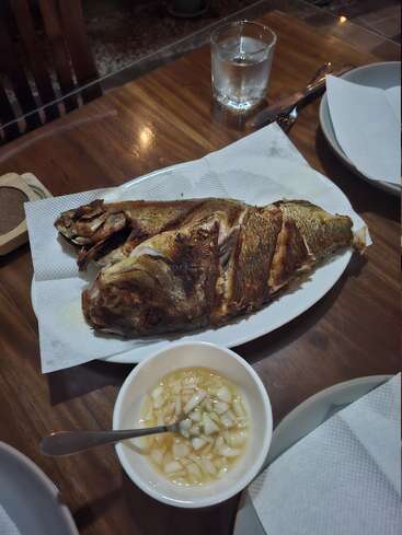 Un poisson entier frit est servi sur une assiette blanche, accompagné d'un bol de vinaigre avec des oignons hachés. Des ustensiles, de l'eau et des assiettes sont également présents.