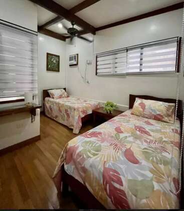 La chambre contient deux lits simples avec une literie colorée à motifs de feuilles, du parquet, de grandes fenêtres avec des stores, un climatiseur mural et une petite table.