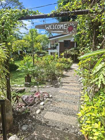 Une allée accueillante bordée de plantes luxuriantes mène à une charmante maison. Un panneau "WELCOME" en bois et des pierres décoratives créent une atmosphère amicale et accueillante.