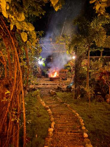 Un chemin de pierre sinueux mène à travers un jardin luxuriant et illuminé jusqu'à un foyer confortable, où les gens se rassemblent, entourés d'arbres et de guirlandes lumineuses.