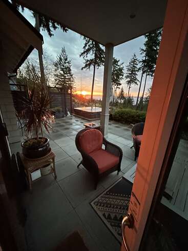 A cozy patio with outdoor chairs, potted plants, and a hot tub overlooks tall trees and a stunning sunset, creating a peaceful and inviting atmosphere.