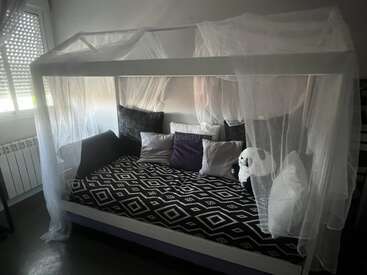 A cozy bed with a house-shaped white frame, draped in sheer curtains. It features multiple throw pillows, geometric patterned bedding, and a stuffed panda.