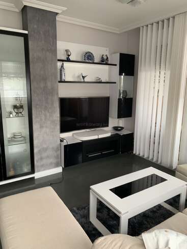 Modern living room with monochrome decor, glass display cabinet, shelves adorned with decorative items, TV mounted on wall, white coffee table, beige sofa, vertical blinds.