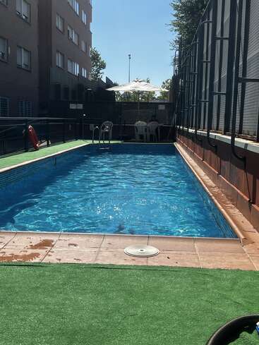 A rectangular swimming pool with clear blue water is surrounded by green artificial grass. There are plastic chairs, a lifebuoy, and a sun umbrella nearby.