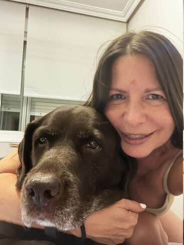 A woman with long dark hair smiles while hugging a large, dark brown dog. They appear indoors with white blinds behind them, radiating warmth and affection.