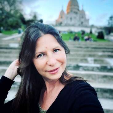Une femme aux longs cheveux bruns prend un selfie, souriante, sur des marches en pierre. À l'arrière-plan, on aperçoit un grand bâtiment en forme de dôme et une pelouse verte.