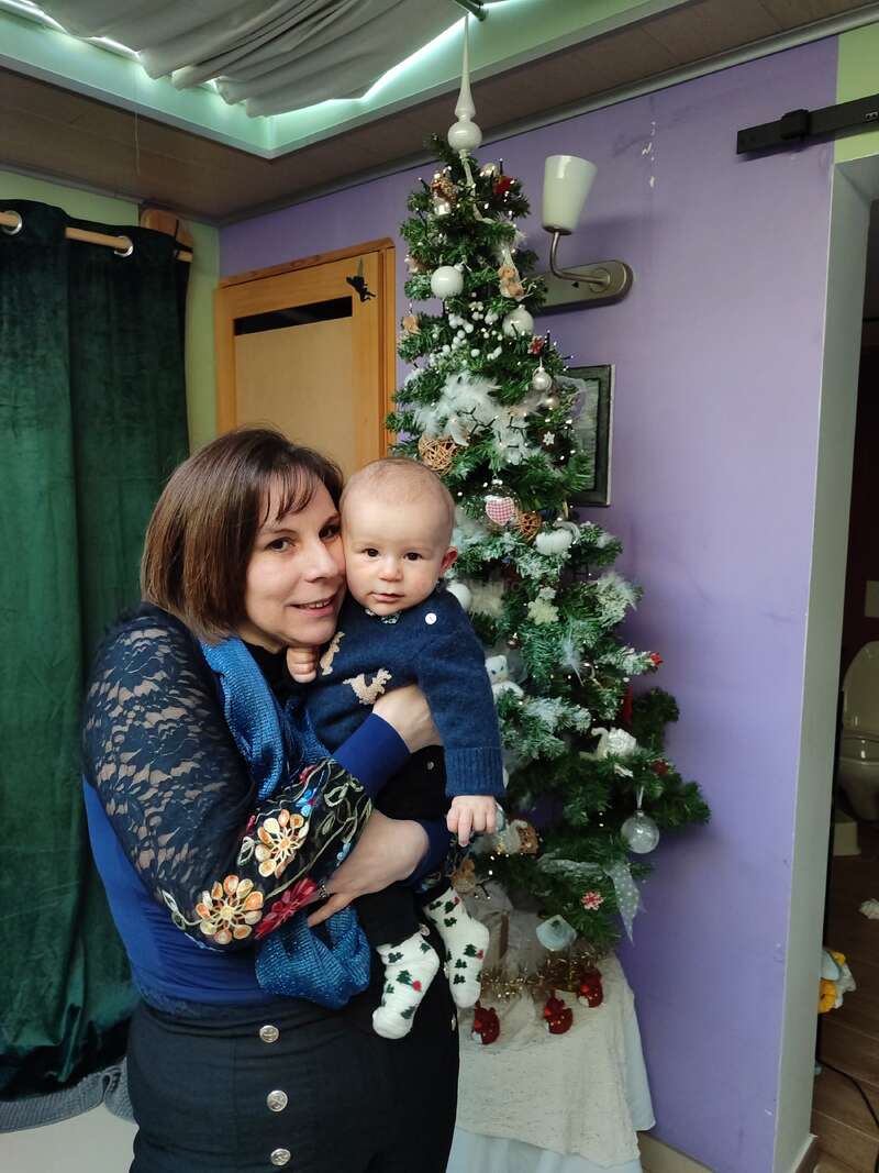Eine Frau hält ein Baby im Arm, beide lächeln sanft, vor einem geschmückten Weihnachtsbaum. Festliche Atmosphäre, gemütlicher Raum, grüne Vorhänge und Weihnachtsschmuck sind zu sehen.