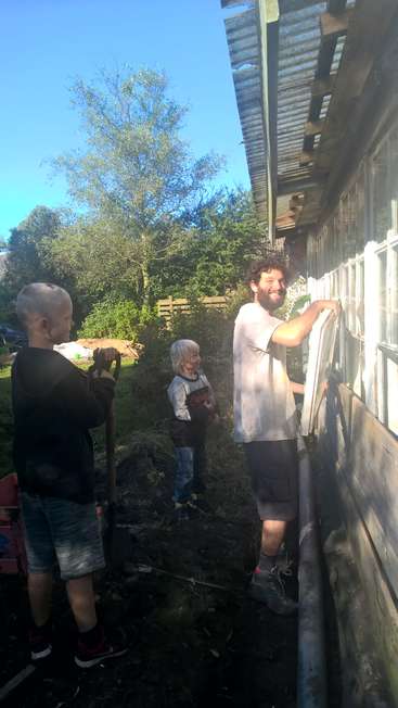 A imagem mostra um homem e duas crianças trabalhando na parte externa de uma casa, com o homem em pé em uma escada e as crianças segurando ferramentas.