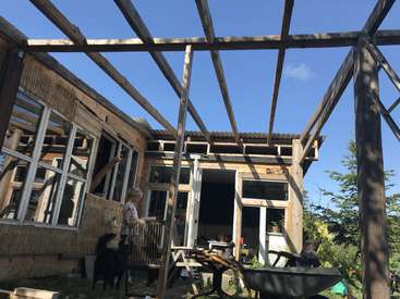A imagem mostra uma mulher em pé na varanda de uma casa em construção, com estrutura de madeira e vigas expostas, cercada por árvores e um céu azul.
