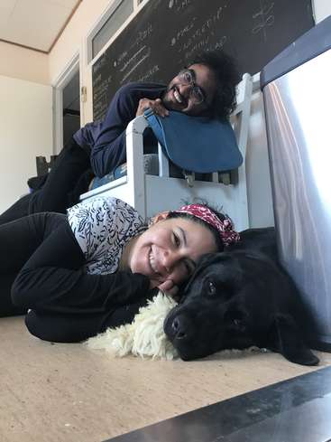 A imagem mostra um homem e uma mulher deitados no chão com um cachorro preto, tendo como pano de fundo um quadro-negro e uma cadeira branca.