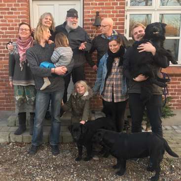 A imagem mostra um grupo de pessoas e cães do lado de fora de uma casa de tijolos, posando para uma foto, com o grupo aparentando ser uma família.