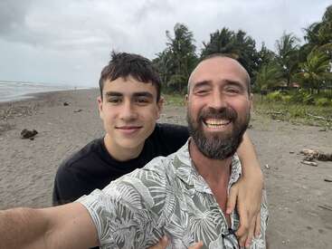 Um cara sorridente e um adolescente tiram uma selfie numa praia tropical, cercados por palmeiras, céu nublado e ondas do mar ao fundo.