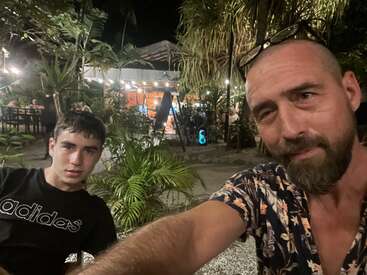 Um cara e um adolescente tiram uma selfie em um restaurante ou café ao ar livre à noite, rodeados por plantas tropicais e luzes ambientais aconchegantes.