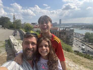 Um cara e duas crianças sorrindo para uma selfie num dia ensolarado, com a cidade, o rio e prédios históricos aparecendo no fundo.