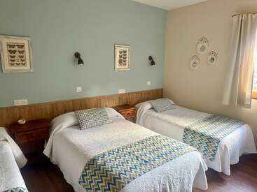 Esse quarto aconchegante tem três camas de solteiro com roupa de cama branca, mantas estampadas, mesinhas de cabeceira pequenas de madeira, quadros com borboletas, pratos decorativos e uma janela com cortina.