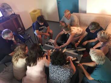 A group of nine people sit closely together in a cozy room, engaging in a board or word game, collaborating, and enjoying each other's company.