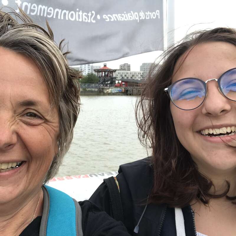 The image shows two women smiling at the camera, with a body of water and a cityscape in the background, possibly taken during the day.